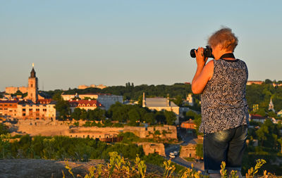 Rear view of female photographer photographing against clear sky during sunset