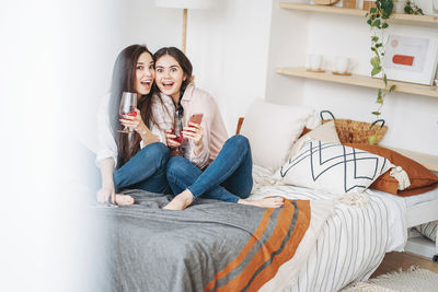 Portrait of female friends having drink while sitting on bed at home