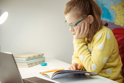 Girl reading book by laptop on table at home