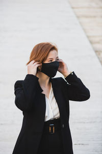 Determined female entrepreneur wearing elegant suit and protective mask standing near building and looking away