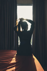 Rear view of young woman adjusting hair while sitting on bed at home