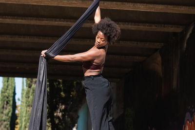 Slender african american female balancing on hanging hammock while doing aerial yoga above ground under bridge in city