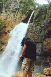 Rear view of man standing by waterfall against mountain