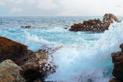 Waves splashing on rocks at shore against sky