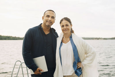 Portrait of smiling mature couple with laptop standing against lake