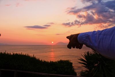 Person hand by sea against sky during sunset