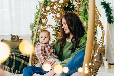 Young  mom is sitting  in a wicker chair with her baby. bokeh of lights of christmas garland 