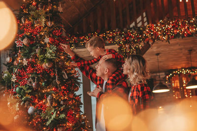 Smile active parents with small son in red checkered sleepwears waiting santa indoor.