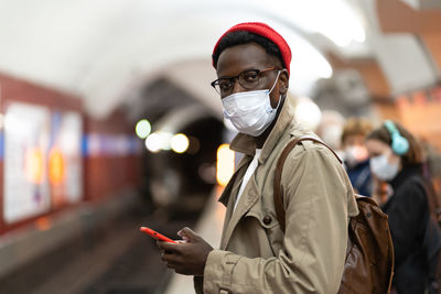 Black millennial man wear face mask as protection against covid-19, virus, waiting train at subway