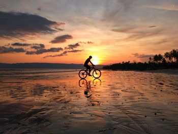 Silhouette person riding bicycle on beach against sky during sunset