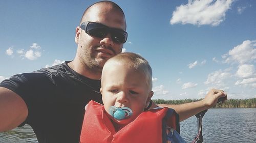 Smiling father in sunglasses with son by lake against sky