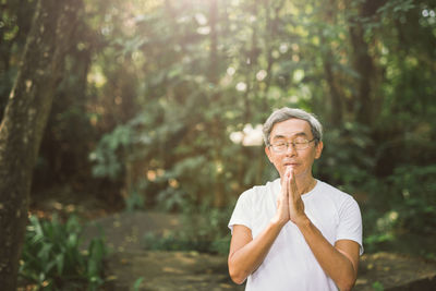 Senior man praying while standing in forest