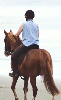 Rear view of person riding brown horse at beach