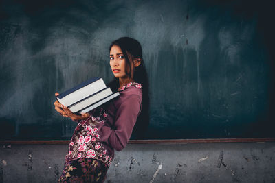 Portrait of young woman carrying books while walking against blackboard in classroom