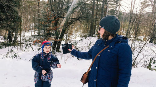 Mother photographing son on snowy field at forest with smart phone