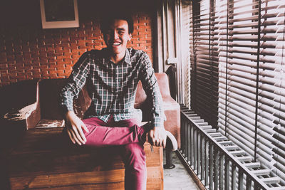 Cheerful young man sitting by window blinds at home