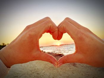 Cropped hands making heart shape at beach against clear sky