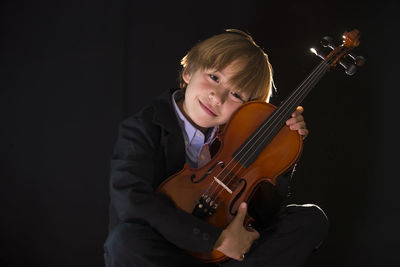 Boy holding violin while sitting against black background