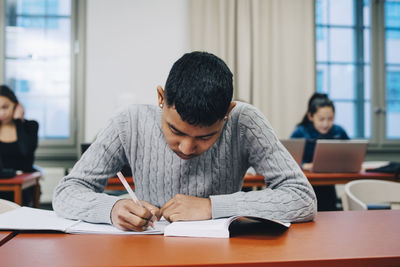 Teenage boy writing on book while studying at desk in high school