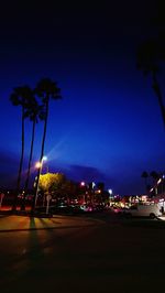 Palm trees by illuminated street against blue sky at night