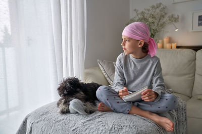 From below happy little child with cancer disease writing notes while sitting with dog on bed in room