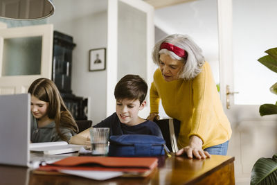 Senior woman leaning on table while assisting grandson doing homework at home