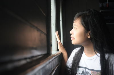 Beautiful young woman looking through window at home