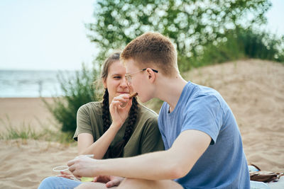 Girlfriend feeding food to boyfriend sitting on sand at beach