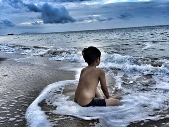 Shirtless boy sitting on shore at beach against dramatic sky