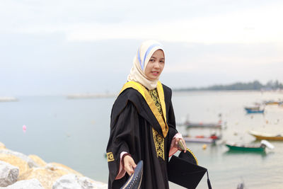 Portrait of young woman in graduation gown standing at beach against cloudy sky