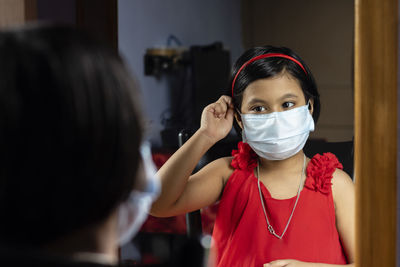 A cute indian girl child in red dress adjusting surgical nose mask in front of mirror