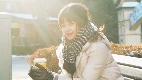 Portrait of young woman holding disposable cup while sitting on bench during winter