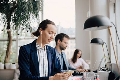 Businesswoman listening music through headphones while sitting with colleagues at desk in office