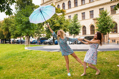 Playful woman holding umbrella while friend pulling hand outside building