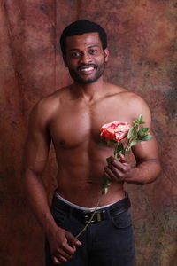 Shirtless young man holding flower while standing against wall