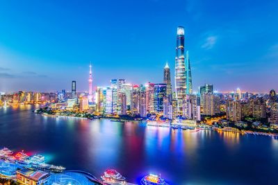 Illuminated buildings by river against blue sky at night