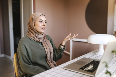 Female freelancer in hijab gesturing during video call through laptop while working at home