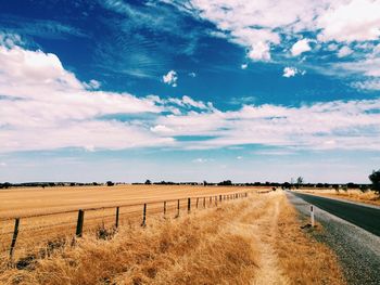 Dirt road passing through field against cloudy sky