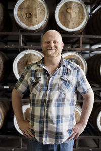 Confident man standing with hands on hip against barrels at brewery