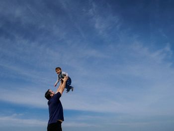 Low angle view of father holding cute baby son while standing against blue sky
