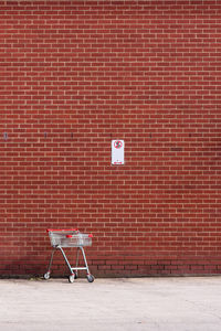 A lonely shopping trolley stays in a non-stoping open space, against a red brick wall.