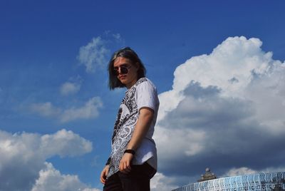 Low angle portrait of teenage boy wearing sunglasses while standing against cloudy sky
