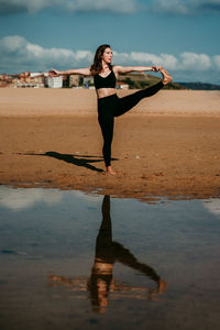 Side view of flexible female standing on sandy shore of river in extended hand to big toe pose and practicing yoga while balancing on leg with eyes closed
