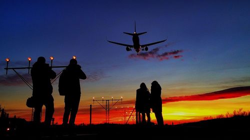 Silhouette people standing against orange sky with airplane