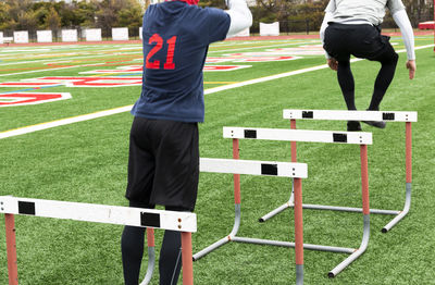 Rear view of two high school boys jumping over track hurdles on a green turf field.