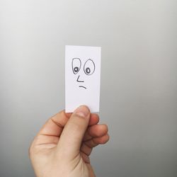 Cropped hand holding anthropomorphic face on paper against gray background