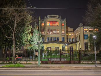 Illuminated street by buildings against sky at night