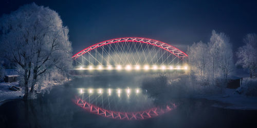 Illuminated ferris wheel by river against sky at night