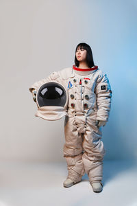 Young asian woman in spacesuit holding helmet and looking away under blue light before mission against gray background