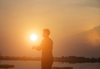 Silhouette man standing by lake against sky during sunset